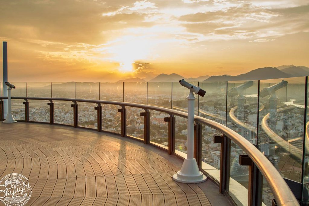 Sunset from the 43rd-floor rooftop at Skylight Nha Trang, with a glass railing and binoculars overlooking the city and mountains